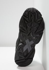 Semelle de chaussure de sport noire avec caoutchouc texturé, présentant un motif de triangles et de crêtes, accent de semelle intermédiaire en gris avec la marque "TORSION".