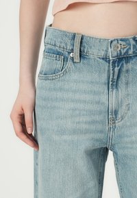 Jean en denim bleu clair avec une taille haute, des poches avant et une texture légèrement délavée. Fermeture à bouton et à glissière avec passants de ceinture.