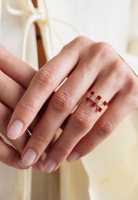 Złoty pierścionek z akcentami w formie czerwonej emalii i małymi przezroczystymi kamieniami szlachetnymi, zaprojektowany w skomplikowany wzór krzyżujących się linii.