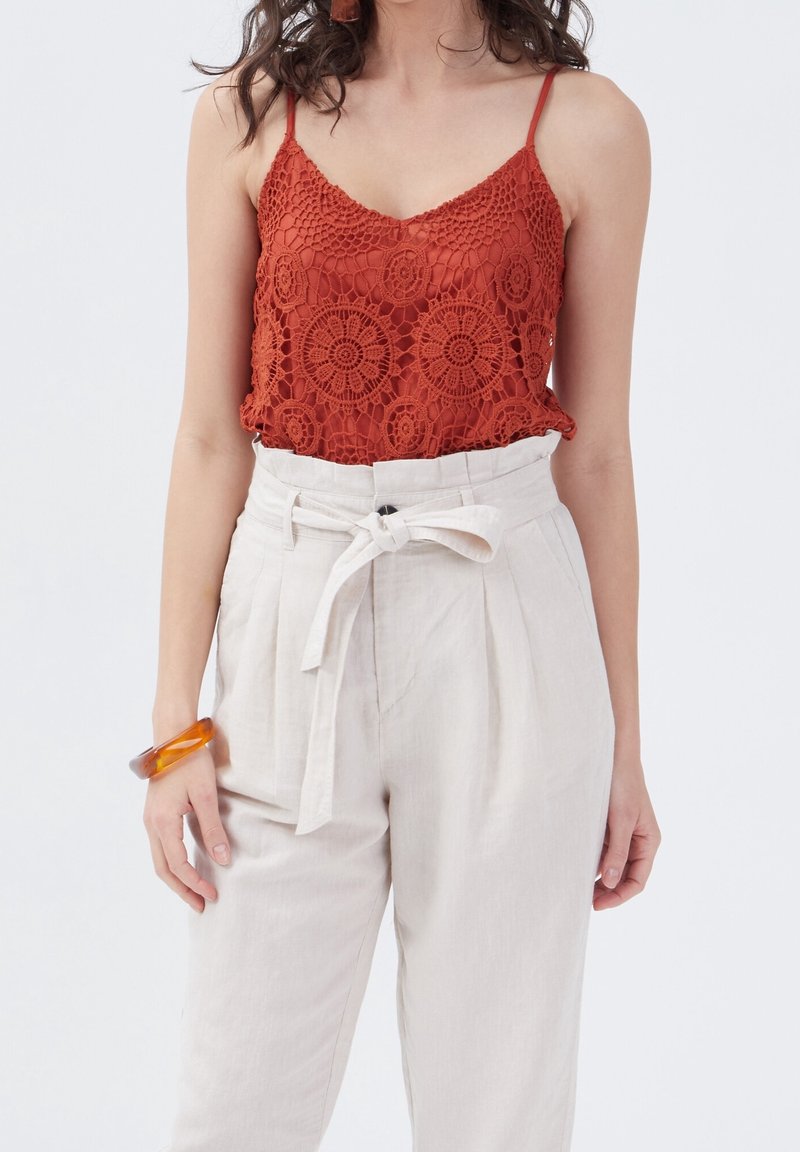 Femme portant un haut sans manches en crochet rouge rouille et un pantalon beige taille haute avec une ceinture nouée et un bracelet en ambre au poignet gauche.
