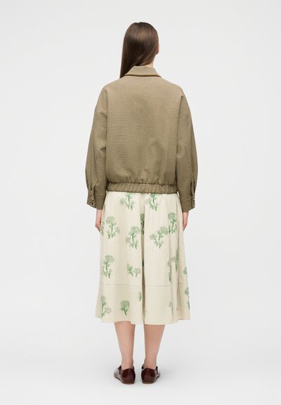 Femme aux longs cheveux bruns portant une veste marron à carreaux et une jupe crème à motifs floraux verts, debout de dos sur un fond blanc.