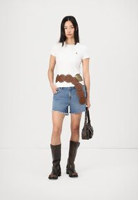 Hvit kortermet t-skjorte, denimshorts, brun skinnbelte med sirkulære detaljer, og knelang brune støvler. Brun håndveske holdt i venstre hånd.