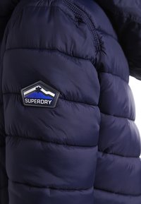 Mörkblå vadderad jacka med quiltad design, med en gummilogotyp placerad på vänster ärm. Slät textur över hela jackan.