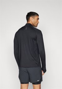 Uomo che indossa una maglia sportiva nera a maniche lunghe e pantaloni corti scuri, in piedi con le spalle alla fotocamera su uno sfondo chiaro e uniforme.