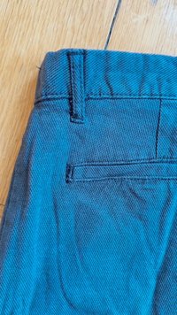 Blauwe textuurstof van een broek met een achterzak, met zichtbare stiksels en een gladde afwerking. De stof heeft een subtiel patroon.