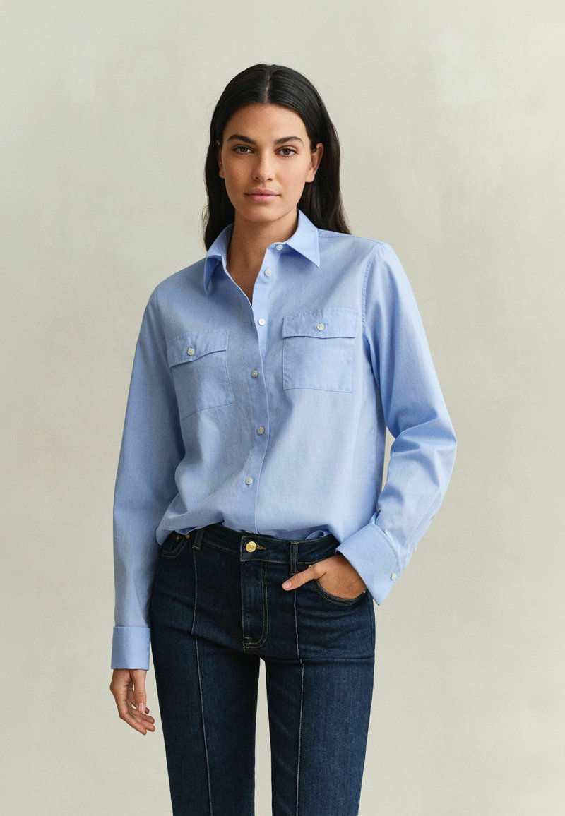 Femme aux longs cheveux foncés porte une chemise bleu clair boutonnée avec des poches poitrine et un jean foncé, debout devant un fond clair uni.