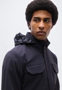 Άνδρας με κοντά σγουρά μαλλιά που φοράει ένα σκούρο μπλε σακάκι με ψηλό γιακά και δύο τσέπες στο στήθος, κοιτάζοντας προς τα αριστερά του.