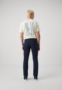 Light beige T-shirt with a large blue "H" on the back, paired with dark navy pants and black shoes featuring white and blue stripes.