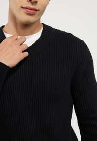 Marineblauer gerippter Strickpullover mit V-Ausschnitt und langen Ärmeln, aus strukturiertem Stoff gefertigt, über einem weißen T-Shirt getragen.
