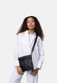 Borsa a tracolla in pelle nera con finitura testurizzata, tracolla regolabile e accessori in oro. Indossata sopra una camicia bianca.