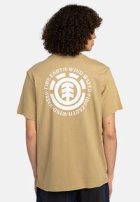 Beige kortærmet t-shirt med et stort hvidt cirkulært grafisk design med elementære symboler på ryggen. Lavet af blødt bomuldsstof.