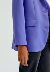 Persona che indossa un blazer viola brillante con dettagli sui bottoni e jeans neri, con la mano rilassata visibile su uno sfondo semplice.