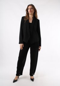 Blazer nero con colletto a scialle, abbinato a pantaloni neri larghi. Stoffa liscia, vestibilità su misura e scarpe nere con tacco alto completano l'outfit.
