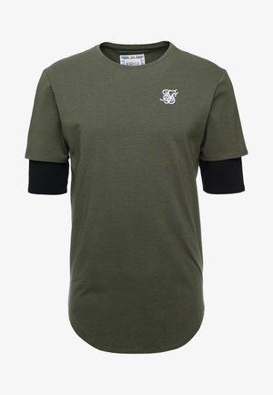 Olivgrünes Kurzarm-T-Shirt, getragen über schwarzen Langarmärmeln, mit weißem, gesticktem Logo auf der linken Brust.