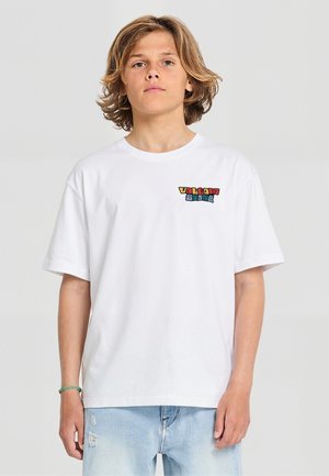 Garçon adolescent aux cheveux mi-longs portant un T-shirt blanc avec un texte coloré "Volcom Stone" et un jean déchiré bleu clair, debout devant un fond uni.