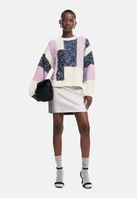 Maglione lavorato a maglia colorato con toppe viola, blu e bianche, con accenti metallici, abbinato a una gonna di jeans argentata e una borsa nera.