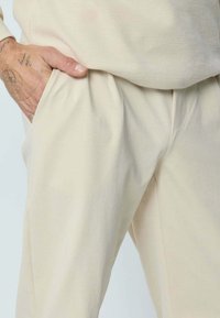 Pantalon en coton beige à coupe décontractée, avec poches latérales, texture douce et détail cousu à la taille pour un look casual.
