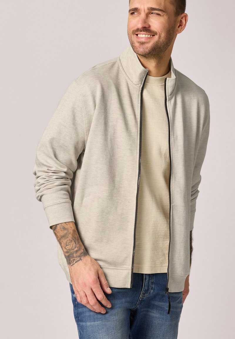 Hombre con chaqueta beige claro con cremallera sobre camisa beige y jeans azules, sonriendo y mirando hacia su derecha, con un tatuaje visible en el antebrazo izquierdo.