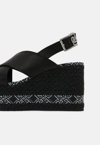 Schwarze Keilsandale mit überkreuzten Lederbändern, silberner Schnalle, geflochtenem Plattformsohle und gemustertem Saum um die Basis.