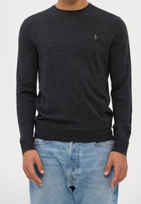 Mu muž nosí tmavě šedý pletený svetr s dlouhým rukávem a vyšitým logem na hrudi, v kombinaci s světle modrými džíny.