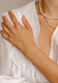 Złota bransoletka z małymi białymi koralikami, z delikatnym zapięciem. Ręka nosząca bransoletkę spoczywa na białym, haftowanym ubraniu.