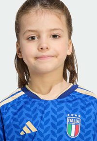 Mladé dievča s hnedými vlasmi, oblečené v modrom futbalovom drese Talianska so zlatými pruhmi a logom, jemne sa usmieva na jednoduchom pozadí.