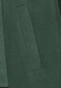 Donkergroene wollen jas met een gladde textuur en een zijzak voor opslag. Het ontwerp heeft een eenvoudig, gestructureerd silhouet.