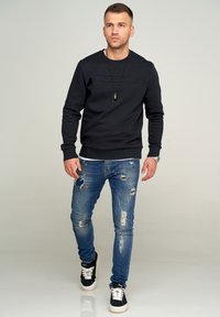 Schwarzer Kapuzenpullover mit strukturiertem Logo, kombiniert mit abgetragenen blauen Jeans. Die Schuhe haben ein schwarzes Obermaterial, weiße Sohlen und schwarze Schnürsenkel.