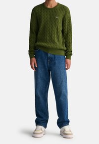 Suéter verde de punto con cable, con puños y bajo acanalados, combinado con jeans azules sueltos de mezclilla y zapatillas beige con detalles en blanco.