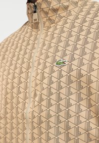 Beige zip-up jakke med et geometrisk mønster i brun, med et grønt Lacoste krokodillelogo på venstre side.