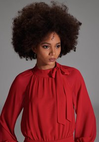 Rode blouse met een hoge halslijn, gerimpelde stof en een gestrikt detail bij de kraag. Doorzichtige stof met lange, vloeiende mouwen.