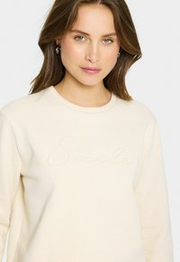 Sudadera color crema con cuello redondo, detalle texturizado de "Smile" en el frente y puños acanalados. Tejido suave con una apariencia acogedora.