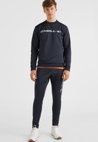 Marineblauw sweatshirt en broek set van zachte stof, met een groot wit logo, oranje accent op de tailleband en een aansluitend ontwerp.