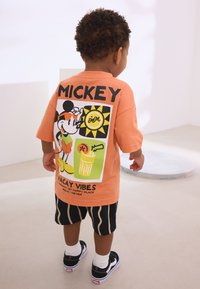 Orange graphic T-shirt featuring Mickey Mouse with a sun design, paired with black striped shorts and black sneakers.