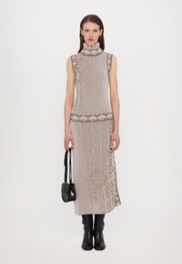 Gilet en tricot sans manches et jupe midi en beige clair avec des motifs sombres complexes en marron. Porté avec des bottes noires jusqu'aux genoux et un petit sac.