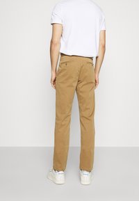 Chinos kaki en coton, avec une coupe droite, des poches arrière avec des boutons, portés avec des baskets blanches.