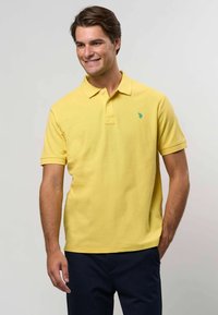 Žluté bavlněné polo tričko s límečkem, dvěma knoflíky a zeleným logem na levé hrudi. Krátké rukávy a mírně uvolněný střih.