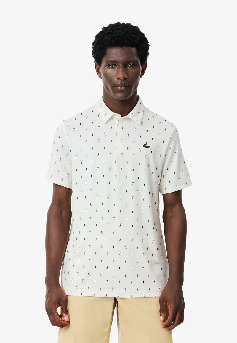 Valkoinen polo-paita, jossa on toistuva tummansininen kuvio, lyhyet hihat, klassinen kaulus ja vihreä logotehoste rinnassa.