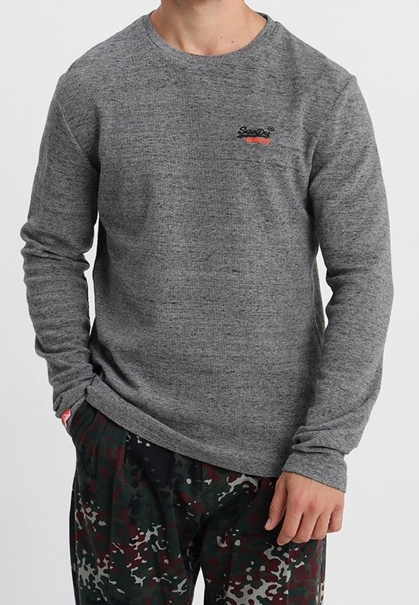 Homme portant un t-shirt gris ajusté à manches longues avec un petit logo sur la poitrine et un pantalon à motif camouflage.