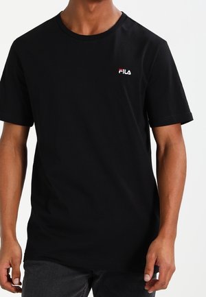 Hombre vestido con una camiseta negra sencilla de FILA de mangas cortas y cuello redondo, combinada con pantalones oscuros, sobre un fondo blanco.