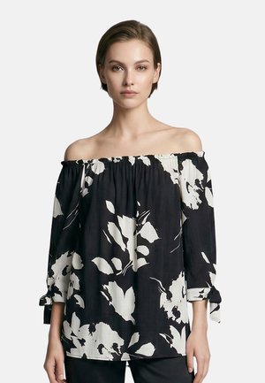 Vrouw draagt een zwart blouse met blote schouders, grote witte bloemenpatronen en driekwart mouwen die aan de manchetten zijn gestrikt.