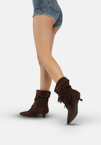 Brązowe zamszowe botki za kostkę z frędzlami, spiczastym noskiem i niskim obcasem; cechują się luźnym krojem i zamkiem błyskawicznym z tyłu.