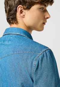 Camicia di denim azzurro chiaro con colletto classico e patta con bottoni, caratterizzata da una consistenza liscia e dettagli di cucitura sottili.