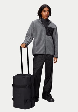 Veste polaire grise à fermeture éclair, poche avant noire, sac de sport à roulettes noir avec des sangles, portant un pantalon et des chaussures noires.