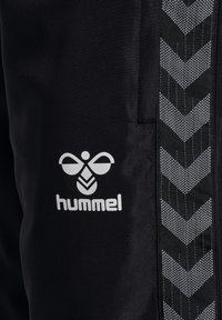 Zwarte sportieve broeken met een textuurstof, wit Hummel-logo en een reflecterende zijstripe met chevronpatronen.