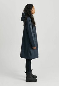 Marineblå vanntett regnjakke med hette, laget av glatt materiale. Har to lommer og sølvfargede trykknapper. Brukt over mørke jeans og svarte støvler.