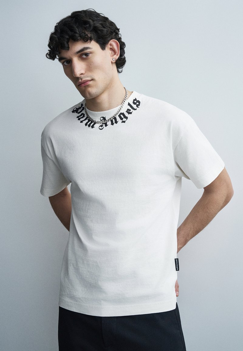 Νεαρός άνδρας με σγουρά σκουρόχρωμα μαλλιά φοράει λευκό T-shirt με μαύρο κείμενο "Palm Angels" γύρω από το γιακά και ασημένιο αλυσιδωτό κολιέ.