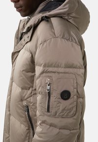 Beige puffer jacket with a textured finish, zippered pockets, and a round logo on the sleeve. Features a hood and a stitched design.