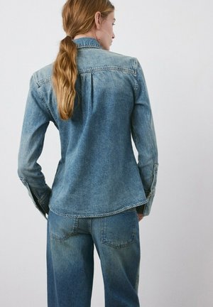 Vrouw met lang lichtbruin haar in een paardenstaart, gekleed in een vervaald blauw denim overhemd en bijpassende spijkerbroek, staand tegen een effen witte achtergrond.
