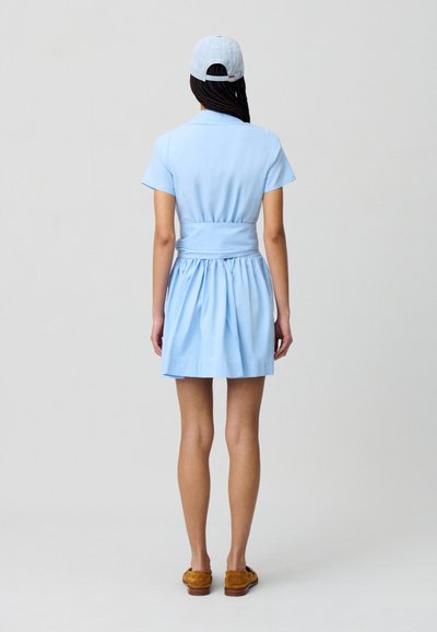 Robe courte bleu clair à manches courtes avec un corsage ajusté et une taille élastique, arborant une jupe évasée. Associée à une casquette bleu clair et des chaussures marron.